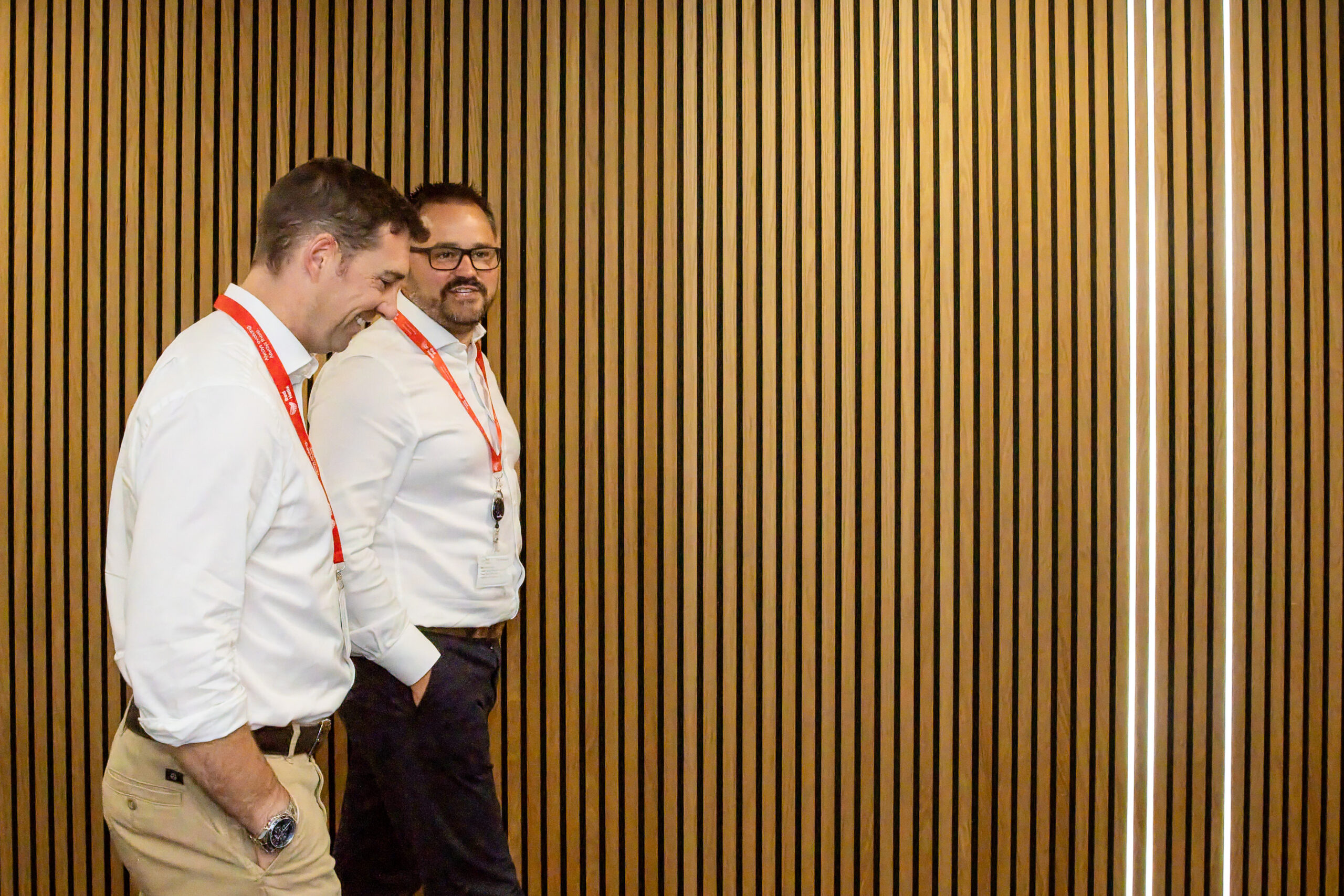 Two men in white shirts walking across a wall panelled with wood